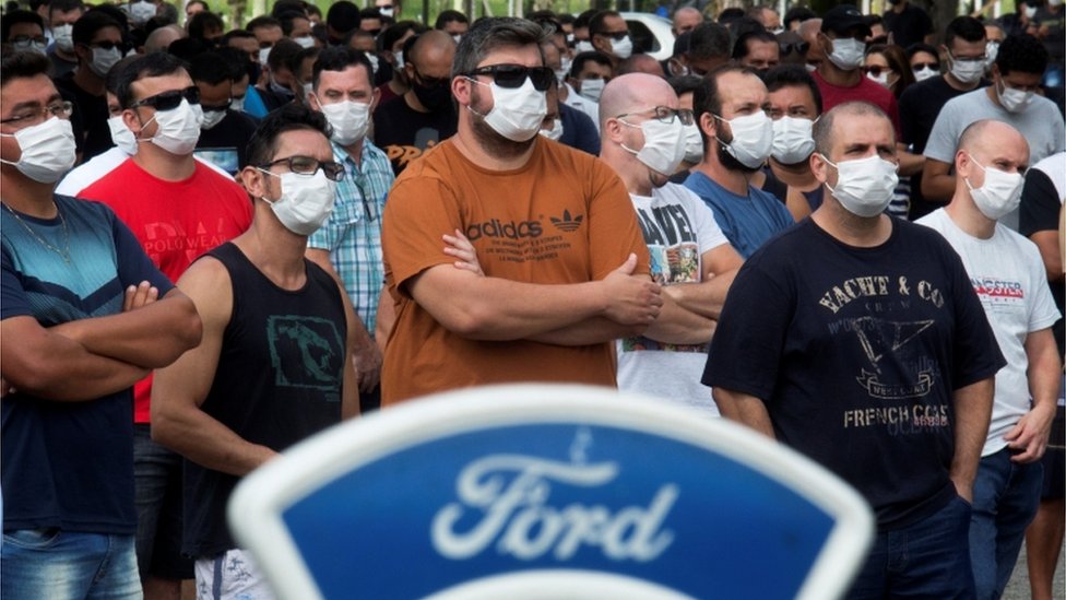 Dezenas de homens e mulheres de máscaras e braços cruzados durante protesto, com logo da Ford na frente destas pessoas
