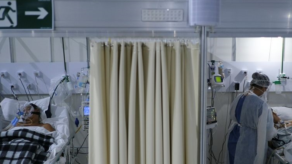 Dois pacientes deitados em macas, separadas por uma barreira; um deles é tocado por uma enfermeira