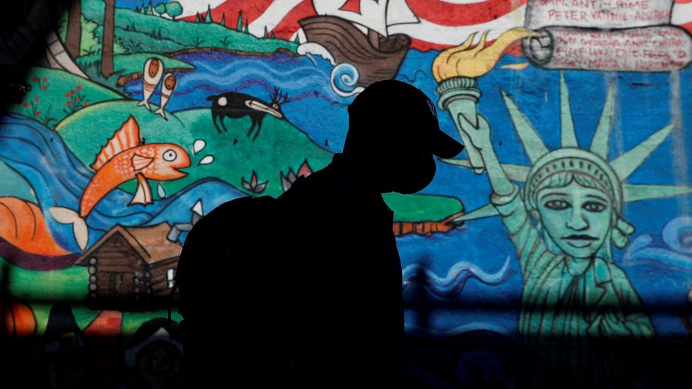 A man in a mask walks in front of a mural in New York