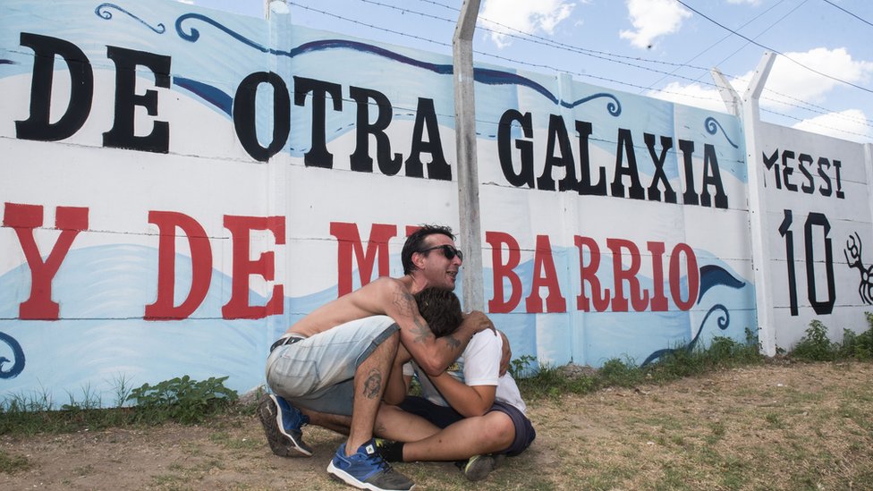 Dos personas se abrazan delante de un mural que dice "De otra galaxia y de mi barrio. Messi 10".