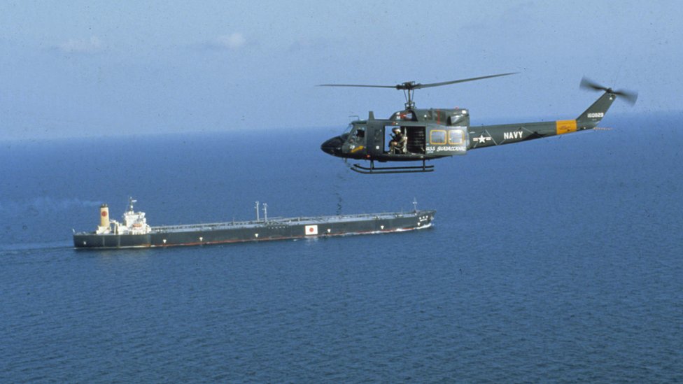 Зеленый вертолет с открытой дверью и видимыми солдатами летит над танкером, несущим что-то вроде японского флага - на хвосте вертолета надпись NAVY и круглые знаки различия ВВС США