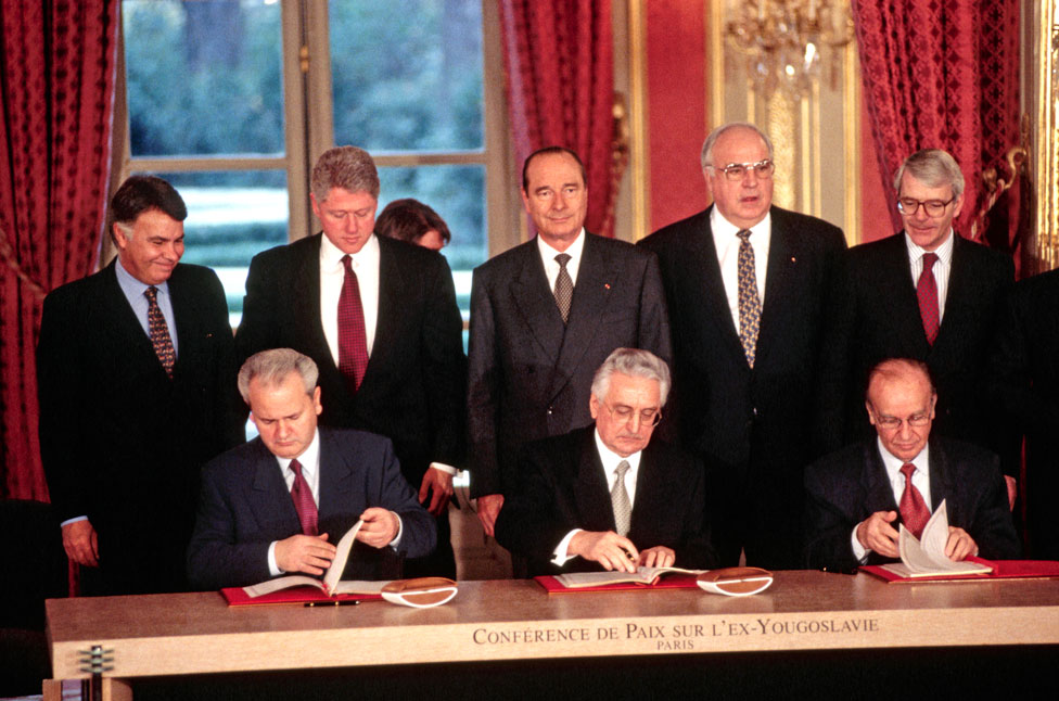 Президенты сидят за столом и подписывают мирное соглашение, а мировые лидеры смотрят сзади