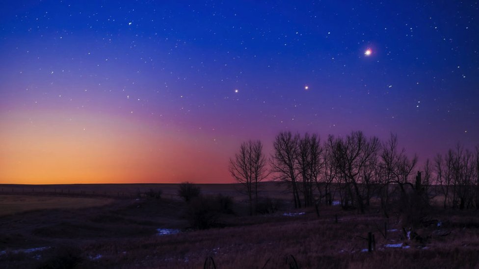 Campo com algumas árvores sob céu ao amanhecer, repleto de estrelas e pontos iluminados