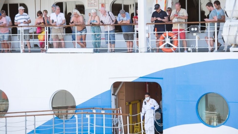 Люди ждут на круизном лайнере AIDAmira, зарегистрированном в Италии, на борту которого находятся в основном немецкие пассажиры, поскольку судно пришвартовано в гавани Кейптауна и находится на карантине из-за нескольких предполагаемых случаев COVID-19 на борту 18 марта 2020 года в Кейптауне Город.