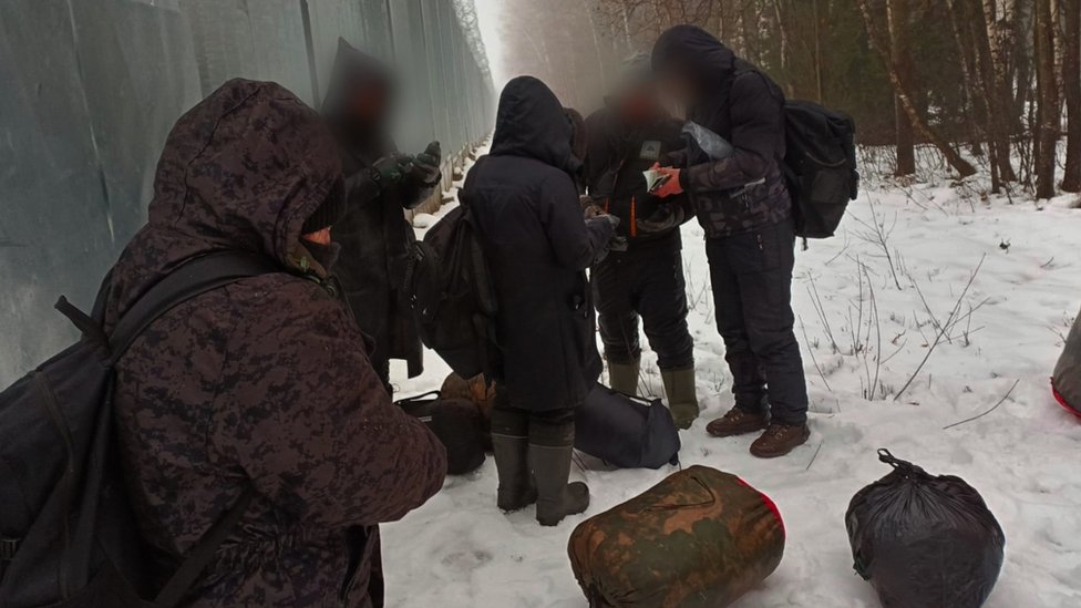 A group of migrants in coats and boots look at their phones in snowy conditions