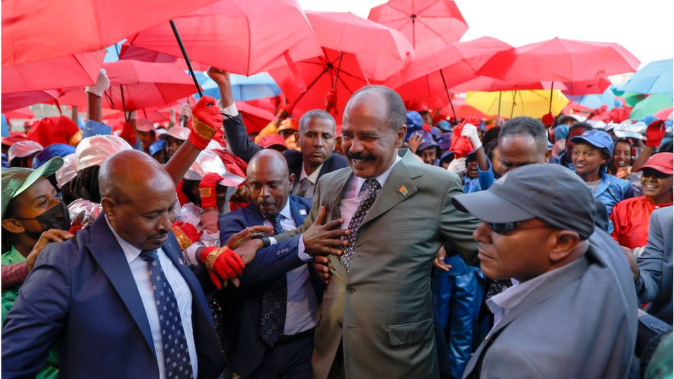 الرئيس الإريتري أسياس أفورقي محاطا بأشخاص يحملون مظلات حمراء. وهو يبتسم.
