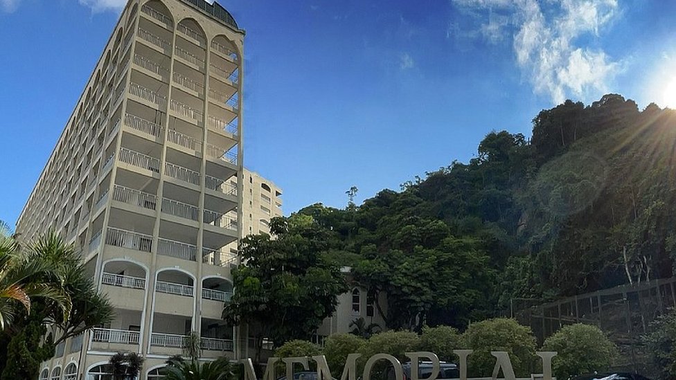 Fotografia colorida mostra um prédio de dez andares sob um céu azul ao lado de árvores verdes