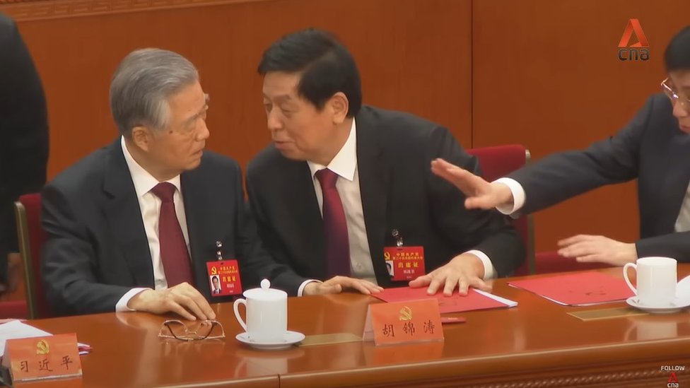 Li Zhanshu (centro) le quita un documento a Hu Jintao (izquierda) mientras Wang Huning (derecha) les hace un gesto.