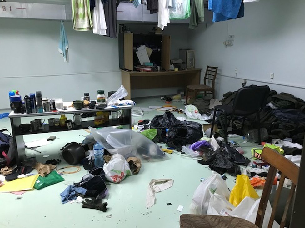 Uma sala caótica com um piso verde pálido coberto com itens como roupas, canecas, cadeiras, sacolas plásticas