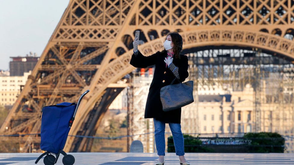 Una turista con mascarilla se toma una foto frente a la Torre Eiffel.