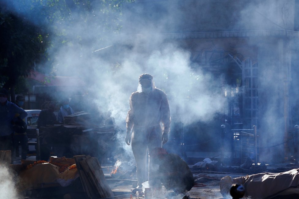 A family member wearing personal protective equipment (PPE) stands next to a body of a person in crematorium as smoke hangs in the air