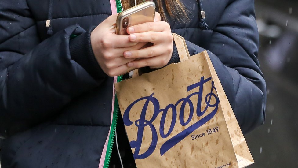 В руках женщины находятся бумажный пакет Boots и мобильный телефон