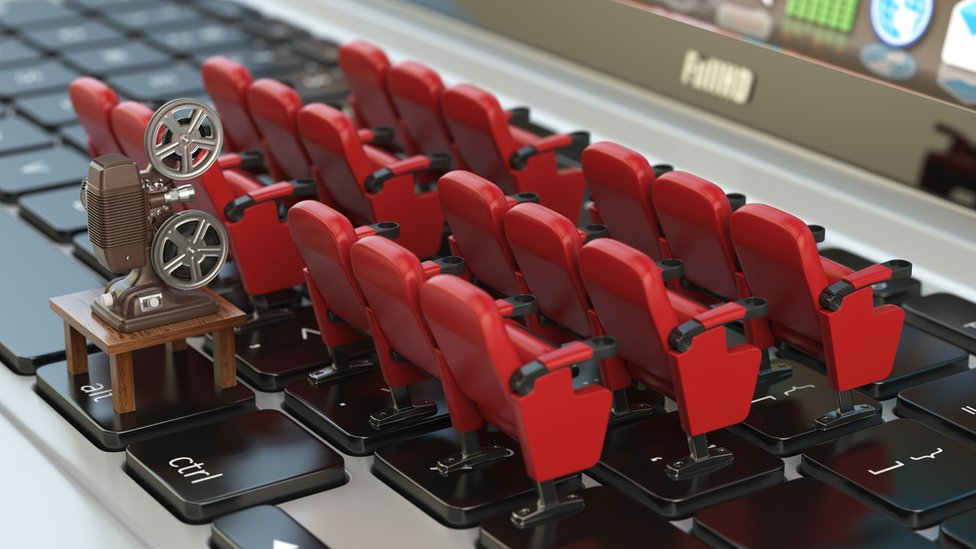 На этой постановочной фотографии модельные сиденья в кинотеатре выстроены аккуратными маленькими рядами на клавиатуре ноутбука, а за ними - модельный кинопроектор в старинном стиле, чтобы проиллюстрировать экран ноутбука, похожий на кинотеатр