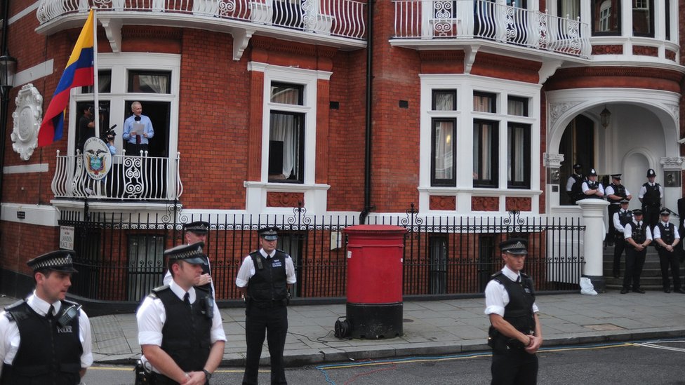 Police officers stand guard as Wikileaks founder Julian Assange addresses the media and his supporters from the balcony of the Ecuadorian Embassy in London, 19 August 2012