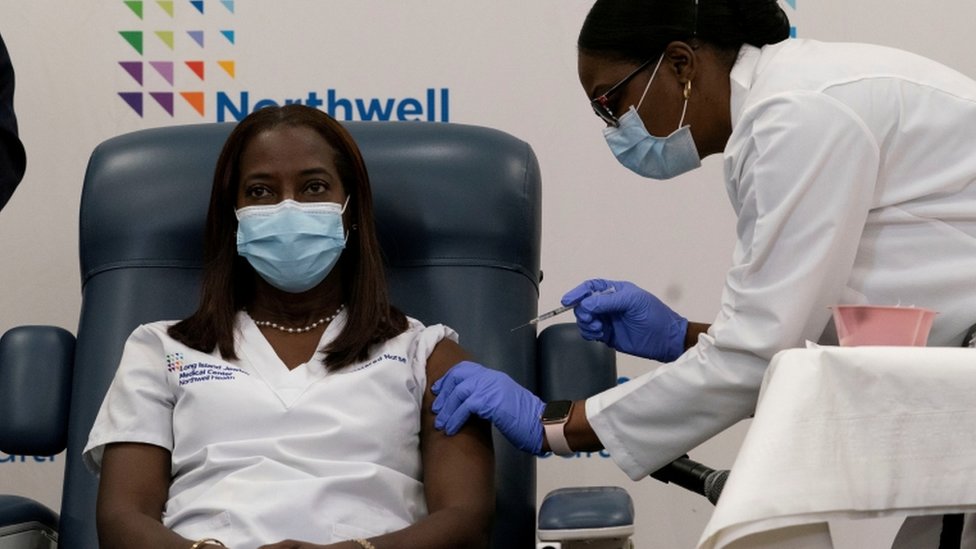 Mulher sentada e de máscara recebe vacina, aplicada por enfermeira, também de máscara e ao seu lado