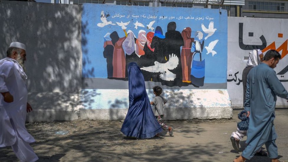 A burqa-clad woman along with a boy walks past a mural depicting women and doves flying along a street in Kabul on September 15, 2021