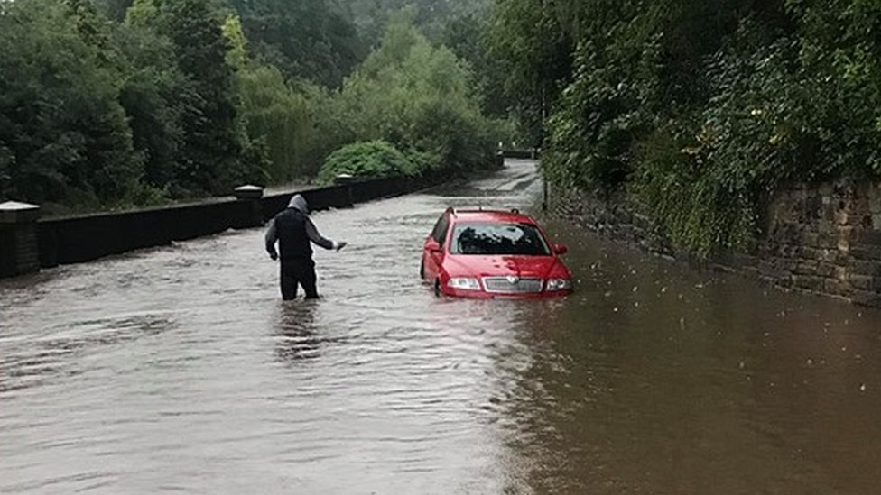 Мужчина пробирается сквозь воду, когда машина застревает на затопленной дороге