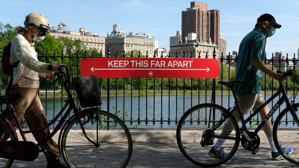 Люди в защитных масках проходят на велосипедах мимо знака социального дистанцирования с надписью «ПРОДОЛЖАЙТЕ ДАЛЬШЕ» у водохранилища Жаклин Кеннеди Онассис в Центральном парке во время пандемии коронавируса 17 мая 2020 года в Нью-Йорке