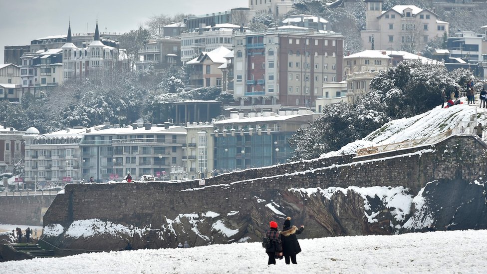 Two people take photos from a white-covered beach with the seaside resort town behind them