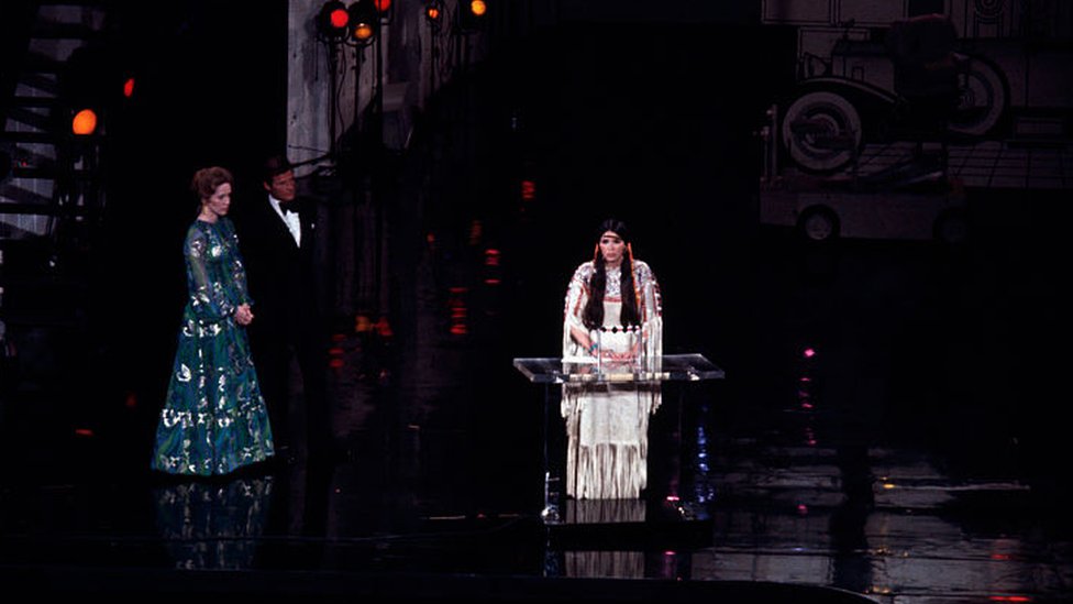 Sobre un gran escenario, a la izquierda, observan Liv Ullman y Roger Moore. Al centro, Sacheen Littlefeather , ataviada con el traje Apache y detrás de un atril, da su breve discurso.