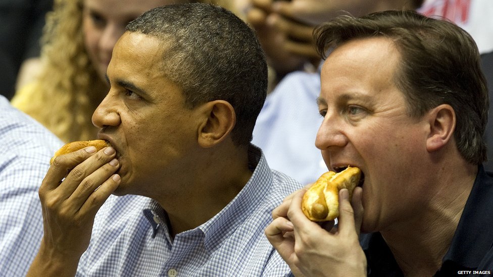 Barack Obama and David Cameron eating hotdogs at a basketball game