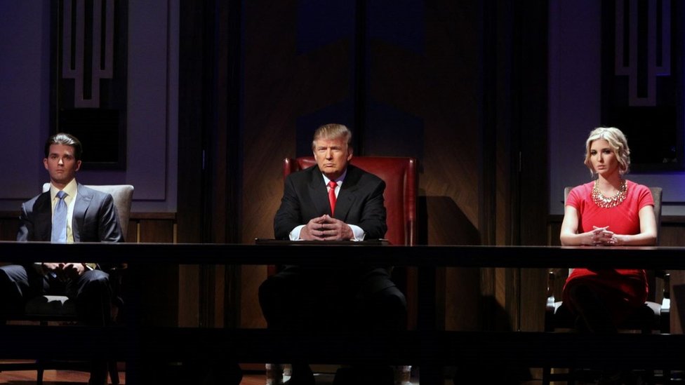Donald Trump on an episode of the Apprentice alongside his two children. All three are sitting behind a large table 