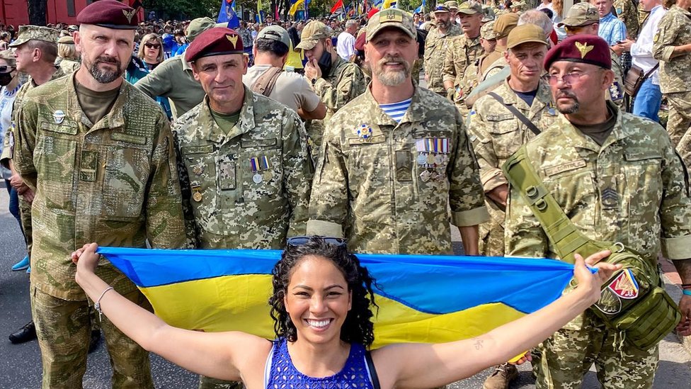 Fernanda Krupin segurando uma bandeira da Ucrânia em frente a homens fardados, durante celebração da independência do país