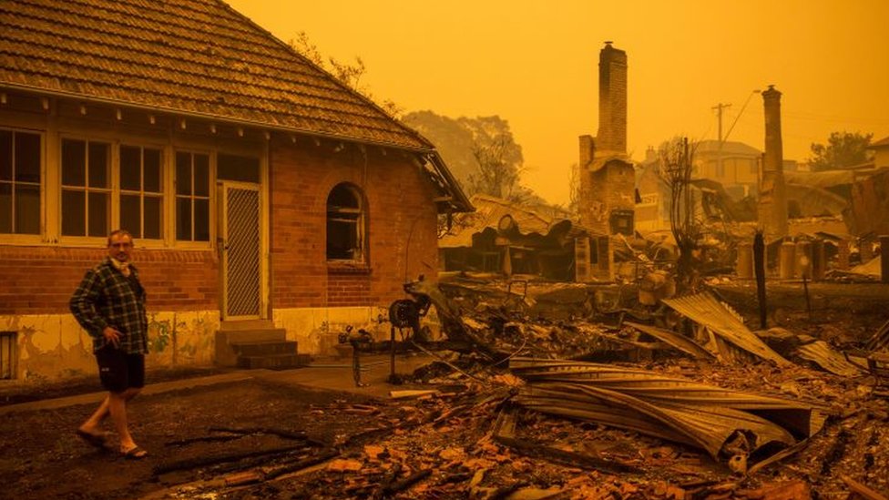 A man stands in front of burnt-out buildings in Cobargo, New South Wales, below an orange sky filled with smoke