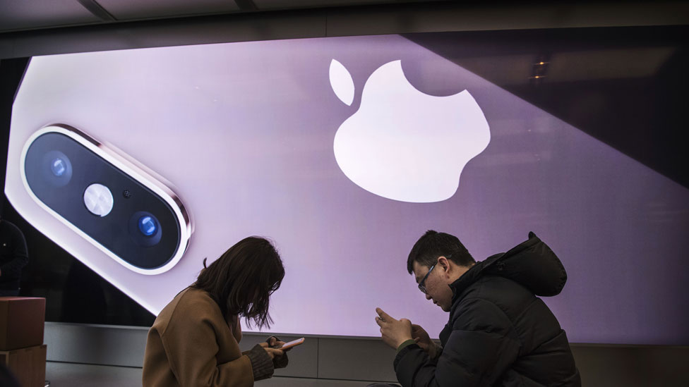 Два человека смотрят на свои айфоны в магазине Apple, и их затмевает огромная реклама последней модели