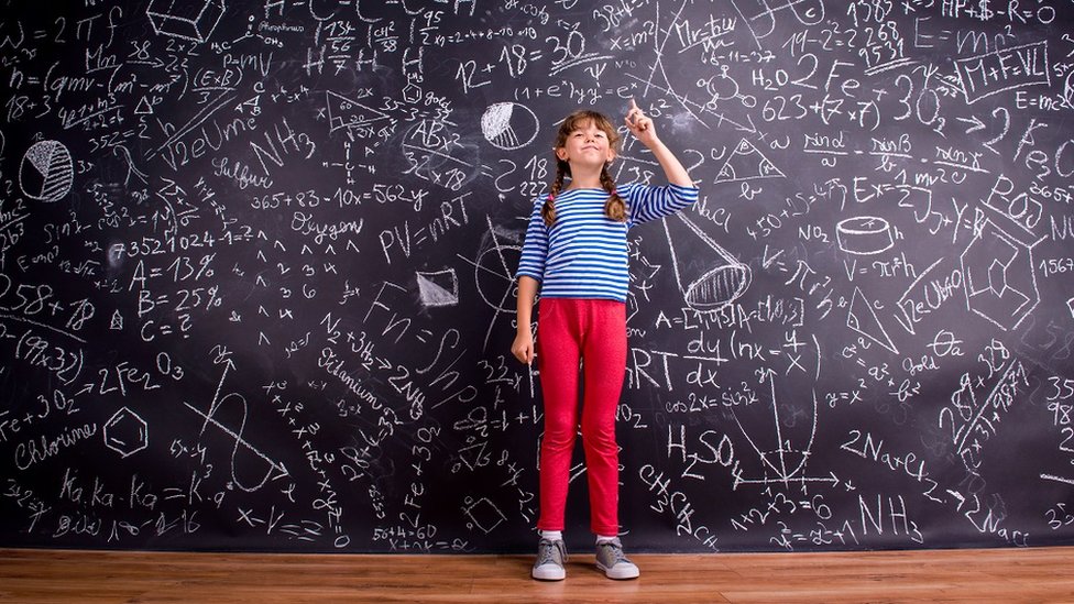 Girl in blue striped t-shirt and red trousers, with two braids, arm raised, finger raised, against big blackboard with mathematical symbols and formulas