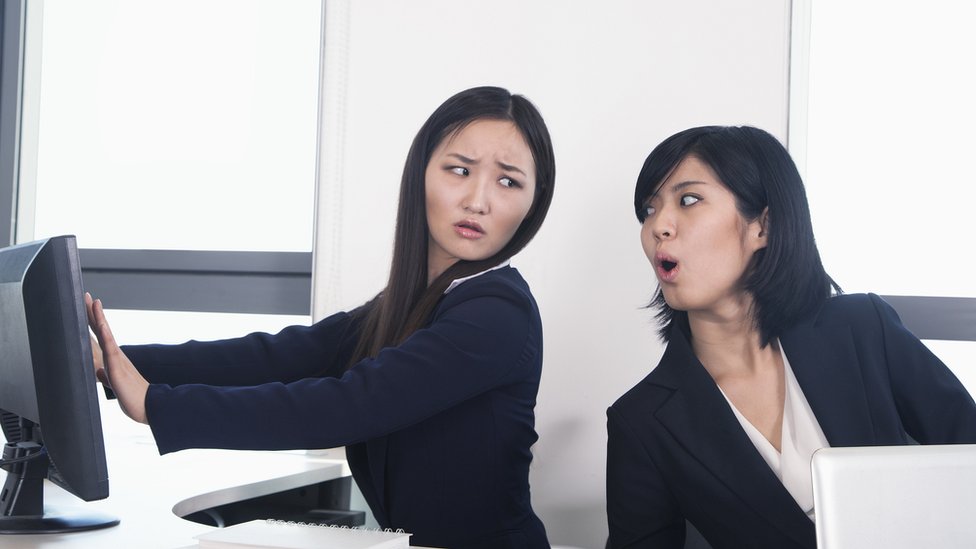 Dos mujeres en la oficina, una de ellas intenta mirar la pantalla de la otra.