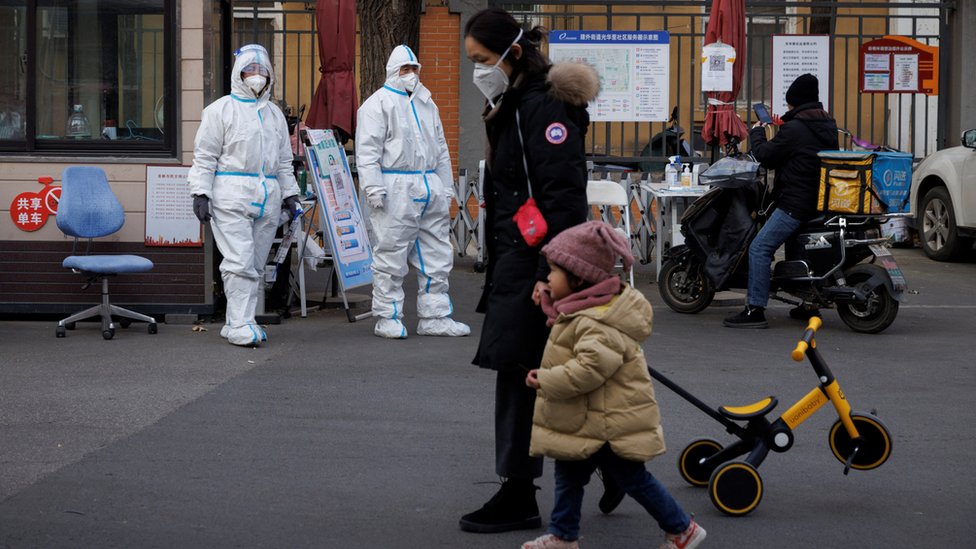 北京某住宅小區外一位女士拖著小孩在兩位身穿防護衣的守衛面前走過（7/12/2022）