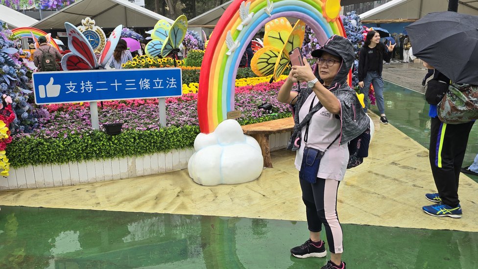 香港花卉展覽上一位女士站在特區政府懲教署園景一面「支持23條立法」標語牌旁拍照（BBC中文圖片18/3/2024）