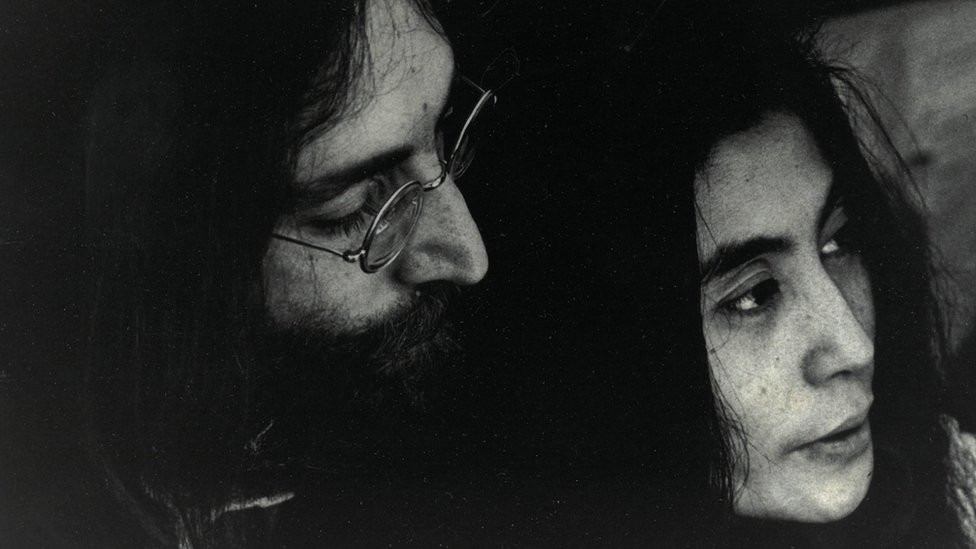 Fotografia em preto e branco em close mostra rosto de John e Yoko, ele está bem próximo à ela