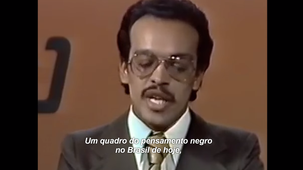 Paulo Roberto Leandro de óculos, bigode fino e terno marrom. Numa legenda em letras brancas, lê-se a fala do jornalista, que diz 