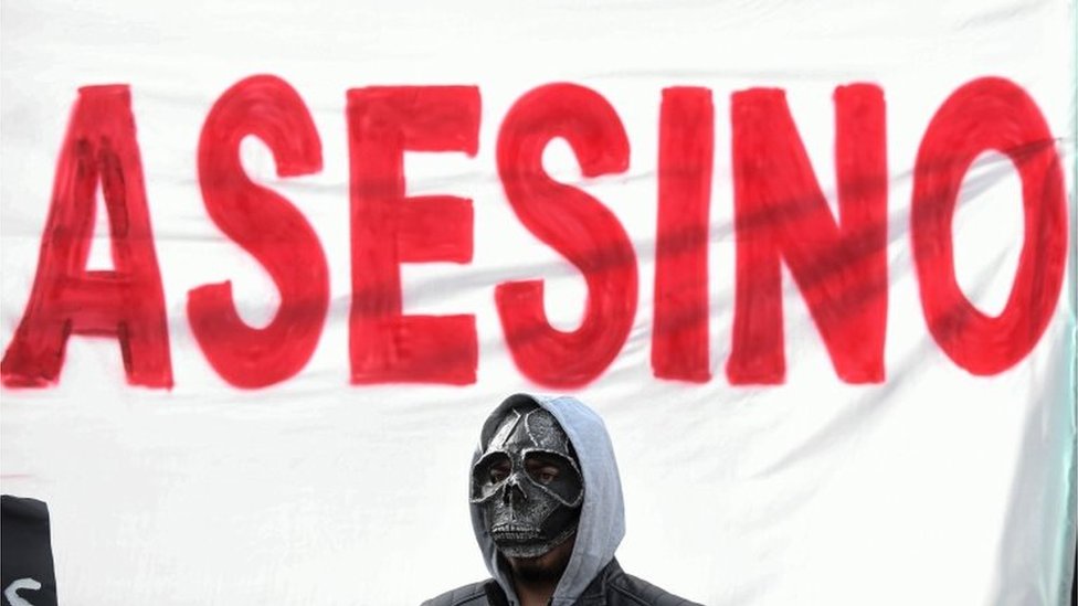 Мужчина в маске стоит перед плакатом с надписью «убийца»