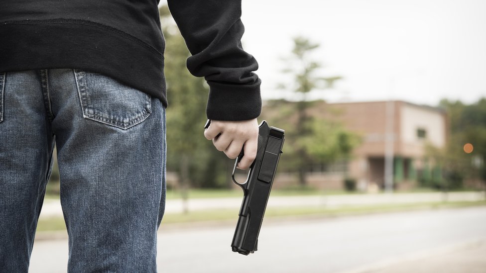 Un adolescente de espaldas con una pistola en la mano, frente a una escuela