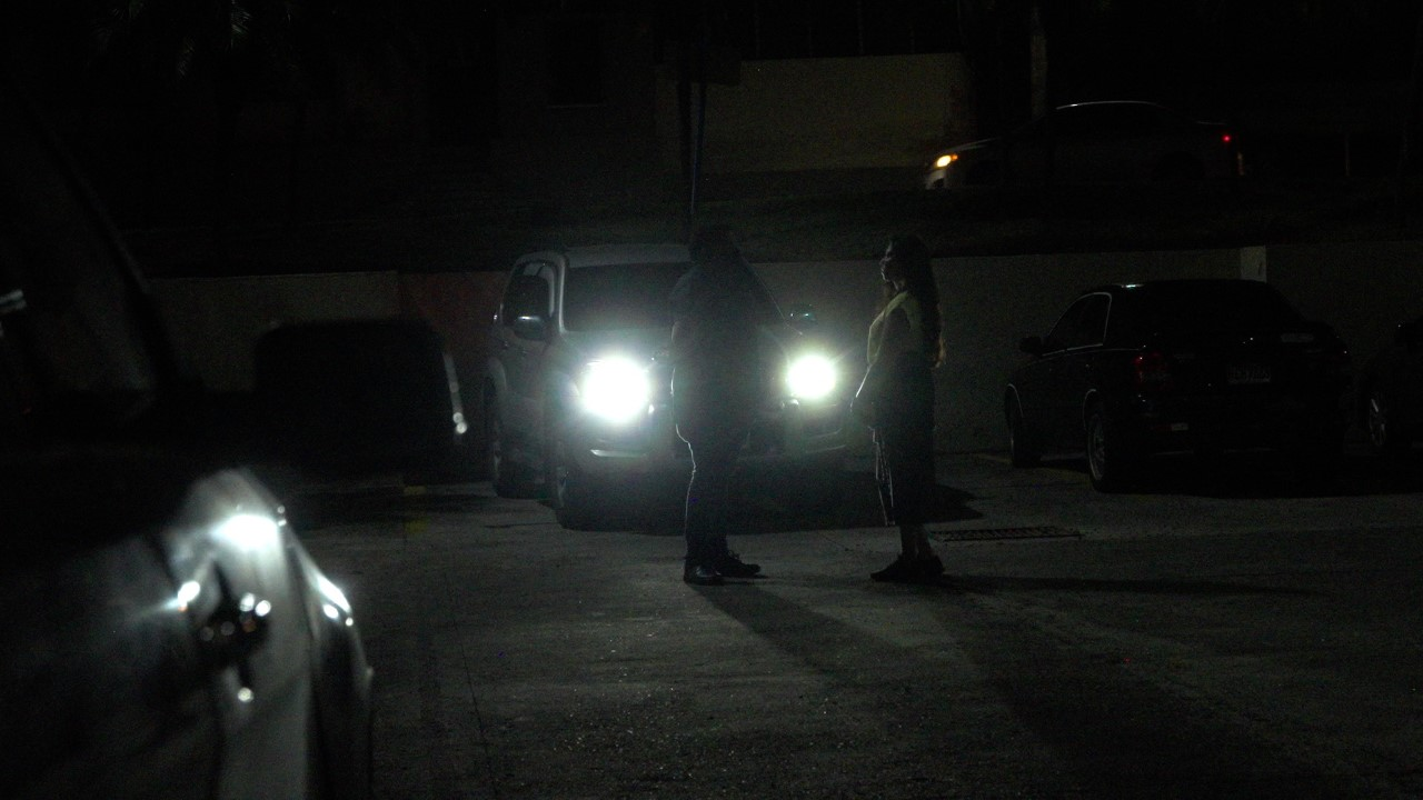 Homem e mulher conversam em frente a um carro com os faróis acesos; é noite e não é possível ver os rostos dos dois