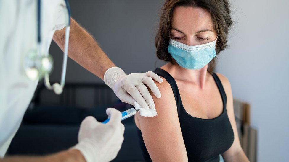 A woman in a mask being injected in the arm looks down at the syringe being held by a masked hand