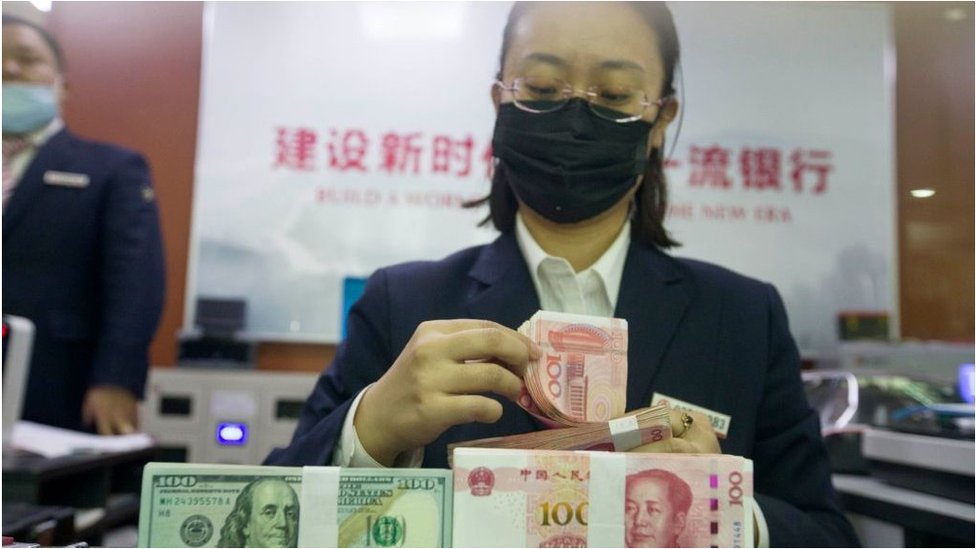 An employee wearing a mask deals with U.S. one-hundred dollar banknotes and Chinese one-hundred yuan banknotes at a bank on October 23, 2020 in Taiyuan, Shanxi Province of China