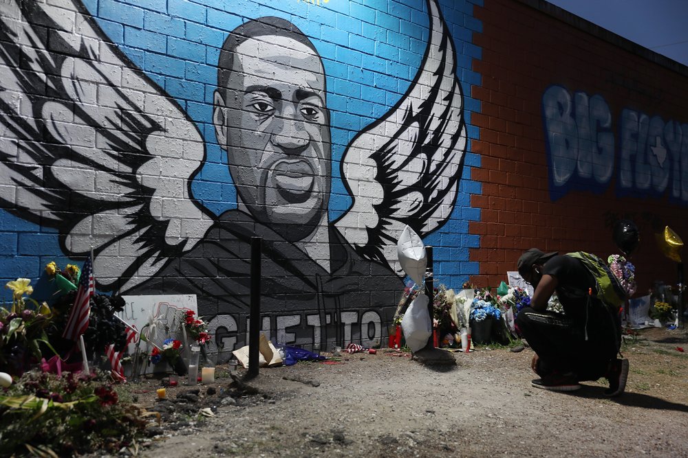 A man kneels in front of a memorial and mural for George Floyd