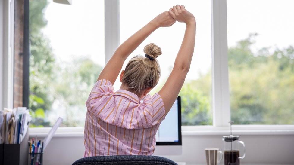 Una mujer de espaldas frente a una pantalla estira sus brazos.