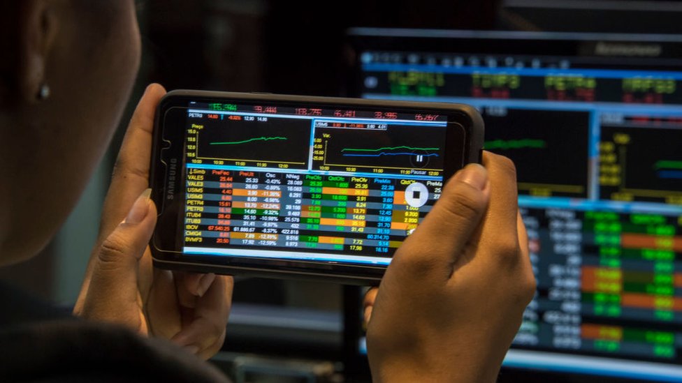 Person with cell phone in front of screen with stock market information in Brazil