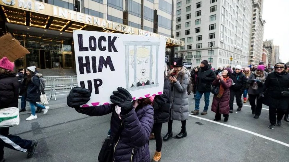 Žena nosi transparent na kome je Tramp nacrtan iza zatvorskih rešetaka