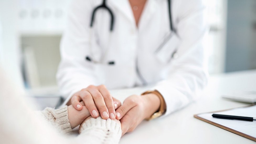 Foto desfocada mostra profissional de jaleco sentado e com mãos em cima da mesa, em um consultório médico