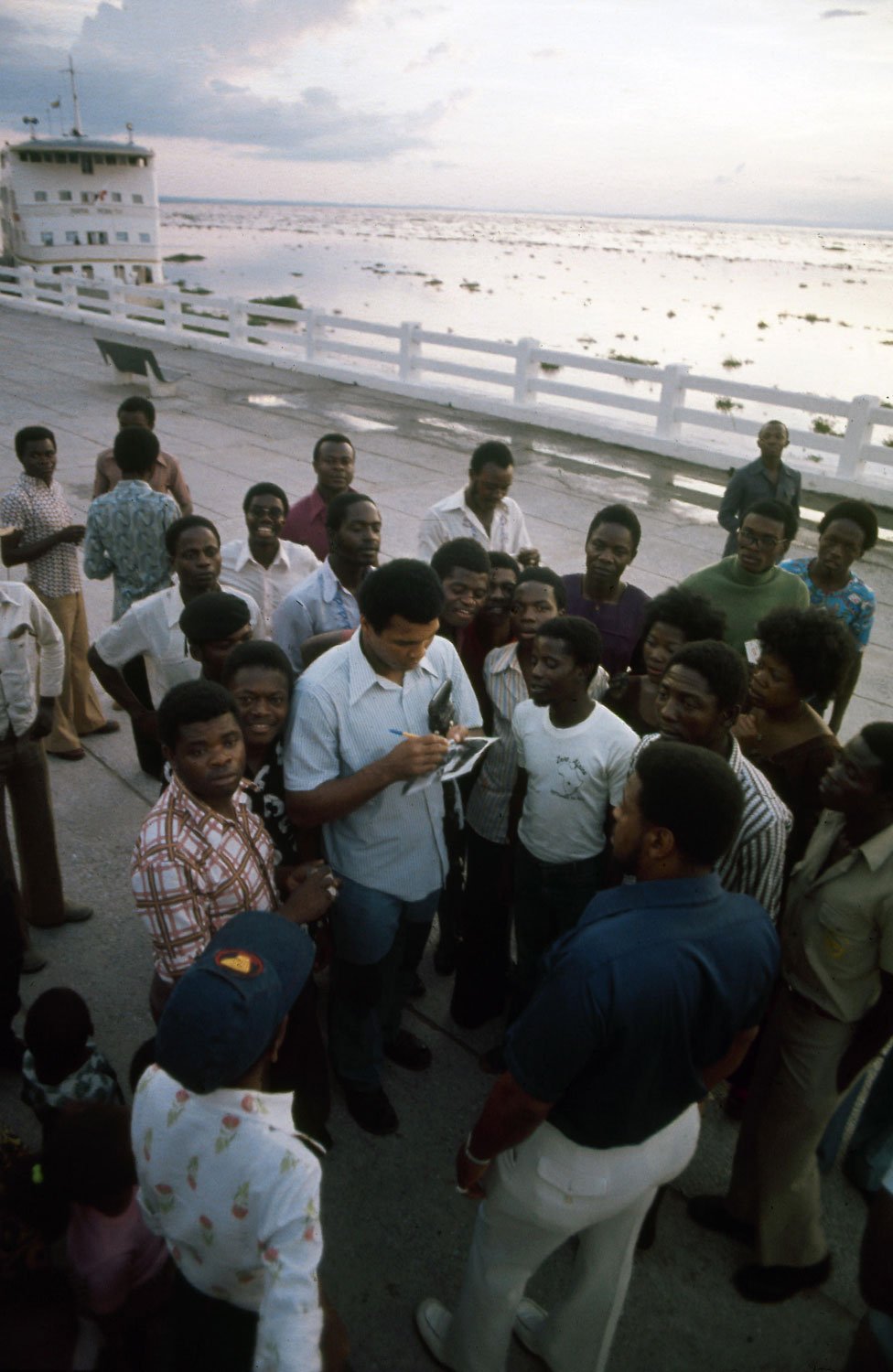 Muhammad Ali signing an autograph amidst a crowd of admirers on the shore of the Congo River at Kinshasa, Zaire, September 1974, prior to his championship fight against George Foreman