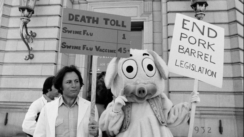 Protesters wearing white coats stand next to a protester in a pig costume, holding a sign saying: "Death toll; swine flu: 1; Swine flu vaccine: 45+" and another saying: "End pork barrel legislation".