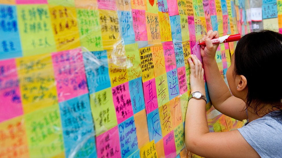 Маленькая девочка пишет на стикере, который нужно повесить на стене, Гонконг, 2014
