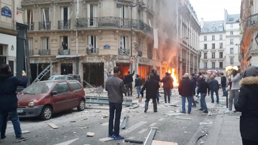 Flames are visible at the site of the bakery as people in the street survey the destruction