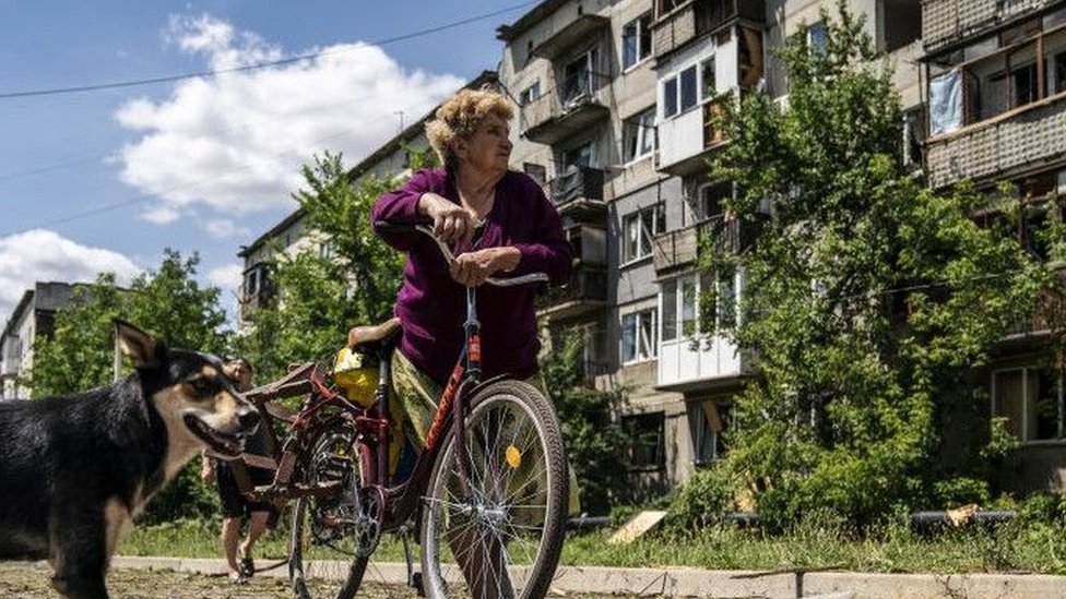 Žena se naslanja na bicikl i gleda u oštećenu zgradu